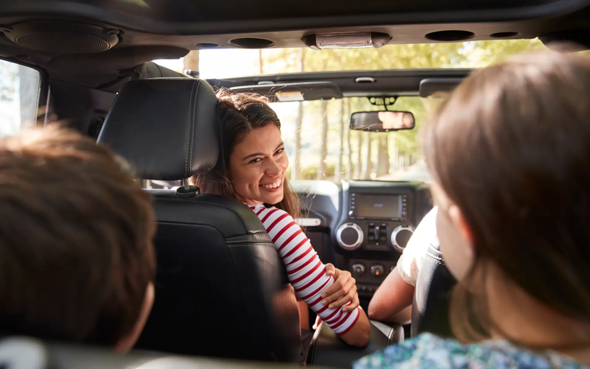 A smiling driver looking back at two passengers in the back of an open-top car on a sunlit road